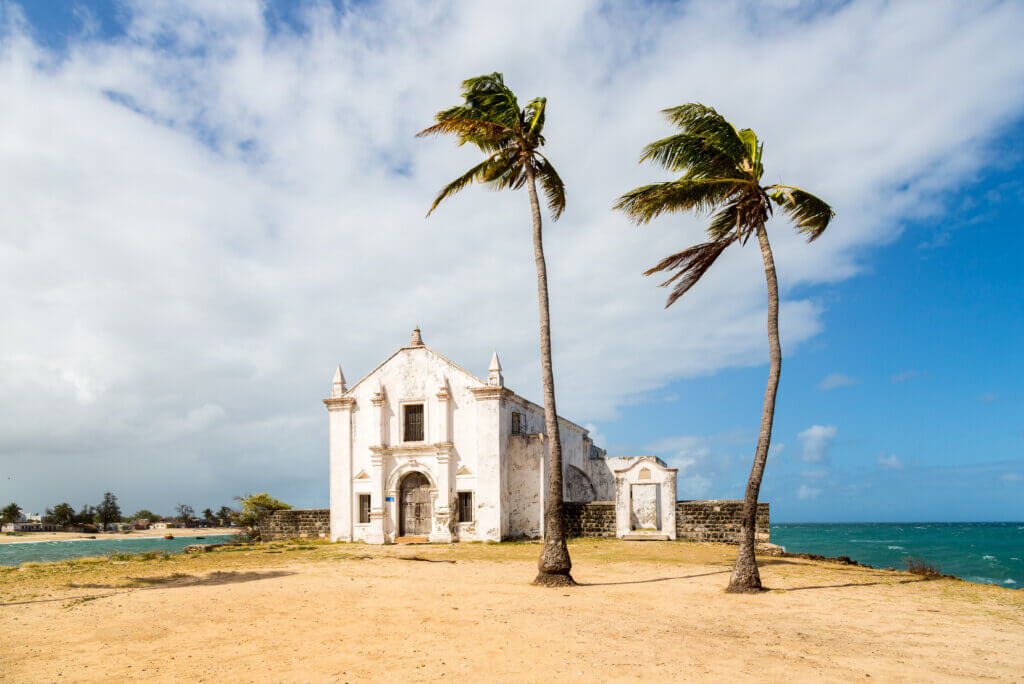 Die Kirche von San Antonio steht auf einer Landzunge auf der Ilha de Mozambique, dahinter strahlt das türkisblaue Wasser des Indischen Ozeans. Vor der Kirche zerzaust der Wind zwei Palmen. 
