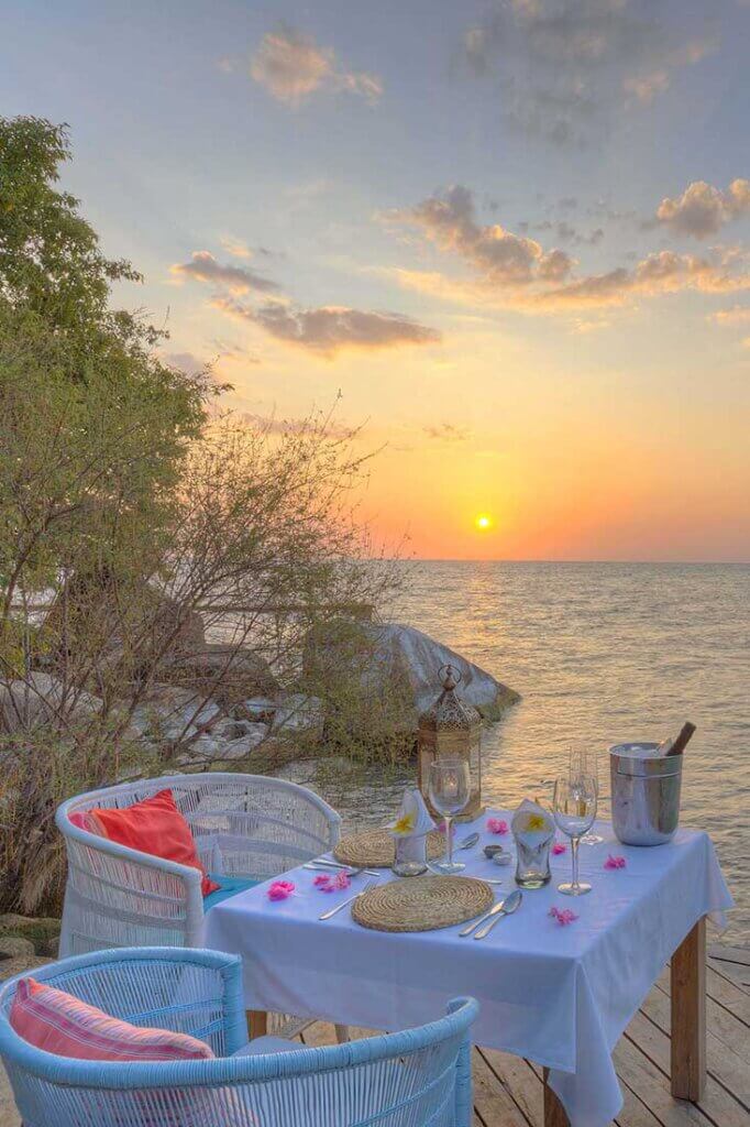 Gedeckter Tisch direkt am Malawi-See bei Sonnenuntergang. Auf dem Tisch steht ein Weinkühler mit Wein, Weingläser, Besteck. Blick auf den Lake Malawi.