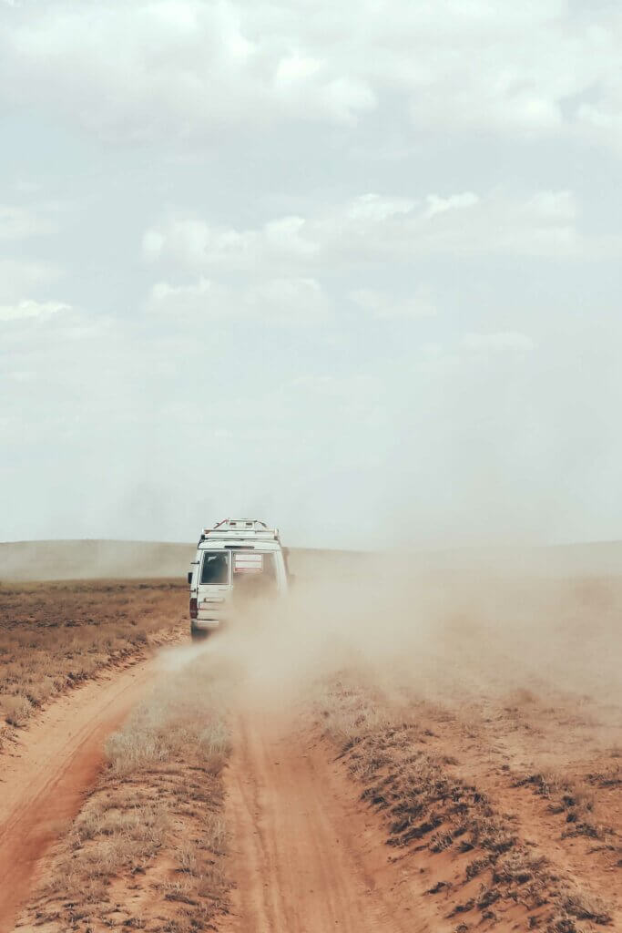 Ein weißer Geländewagen braust in einer verdorrten Landschaft in Afrika auf einer Staubpiste und hinterlässt dicke Staubschwaden. 