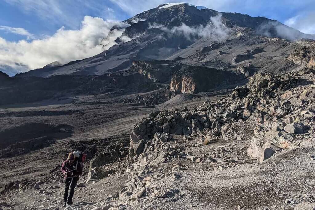 Ein Wanderer steigt vom Kilimanjaro ab. Im Hintergrund sieht man den verschneiten Uhuru Peak, der Wanderer läuft durch Geröllfelder und karges Gebiet.