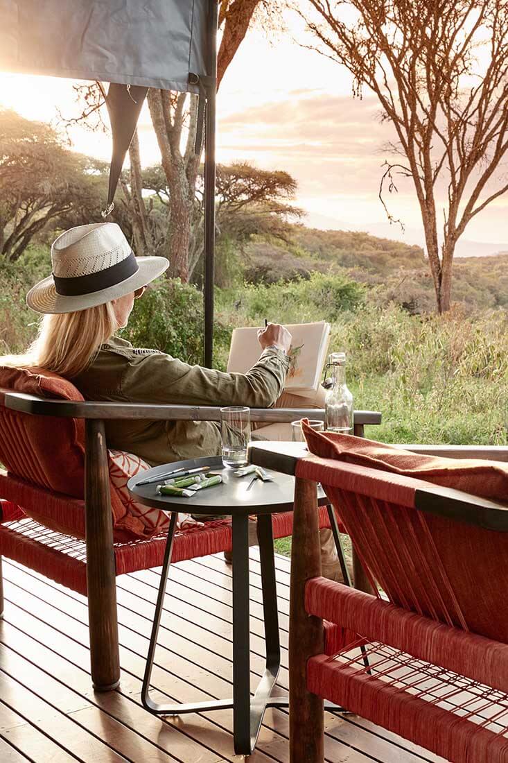 Eine Frau mit Safari-Hut sitzt auf der Terrasse des Sanctuary Ngorongoro Camps und malt.