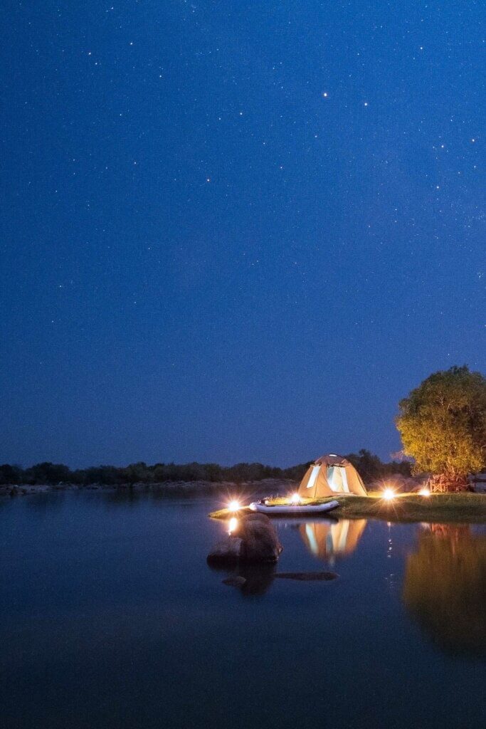 Am Kafue River steht ein hell erleuchtetes Kuppelzelt unter dem Sternenhimmel. Das Zelt wird als Unterbringung für ein mehrtägiges Kanu-Abenteuer verwendet. 