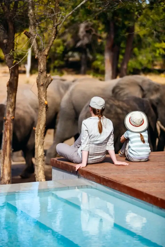Eine Mutter und ihre Tochter sitzen am Pool und beobachten eine Herde Elefanten. 
